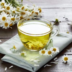A chamomile herbal tea with flower buds nearby on a wooden table with a textile and a camomile bouquet